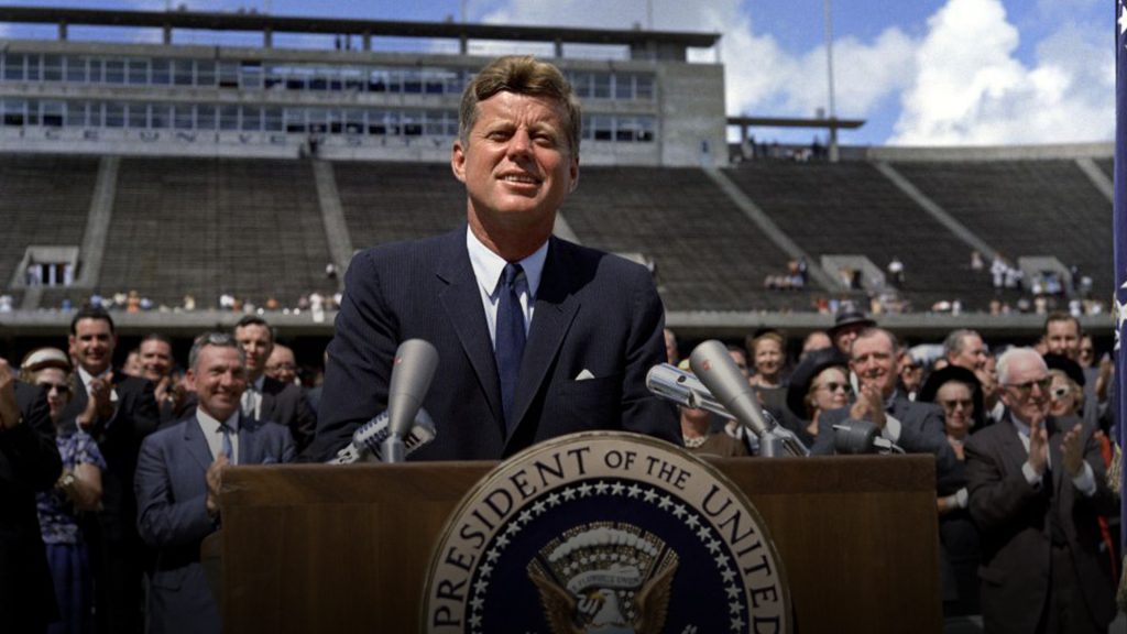 President John F. Kennedy delivering his famous 'We choose to go to the Moon' speech at Rice University stadium in 1962, standing at a podium before a large crowd.